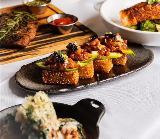 An elegant plate of crispy rice topped with avocado and a savory topping is displayed on a table, surrounded by dishes of grilled steak, salmon with vegetables, and a side dish in the foreground.