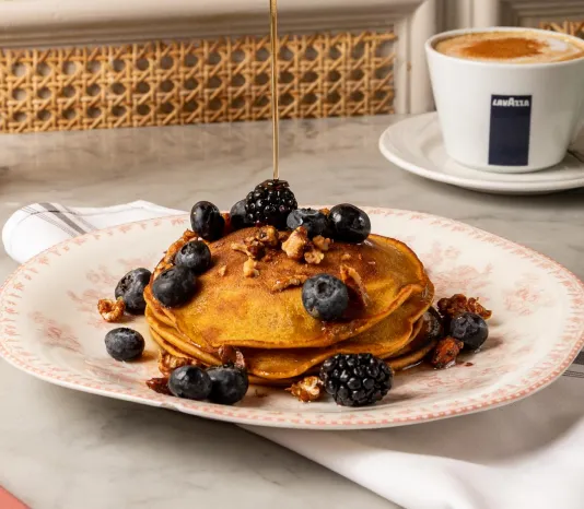 A hand pours syrup onto a stack of pancakes topped with blueberries, blackberries, and nuts; a cup of coffee sits in the background.